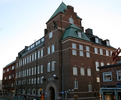 Tegel hus i flera båningar med grönt plåttak med takkupor. Fönster med flera rutor och vita spröjs.