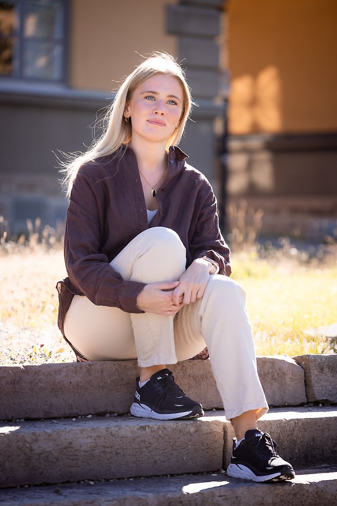 En yngre kvinna med ljust långt hår, mörk jacka och ljusa byxor sitter på en stentrappa i solen