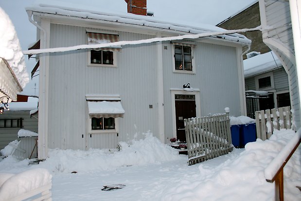 Trähus i två våningar i ljusgrå stående panel med vita knutar och detaljer. Fönster med svarta spröjs och svart ytterdörr.