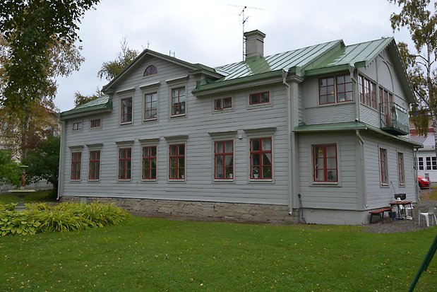 Trähus i trevåningar i ljusgrå, liggande panel, med grå knutar, fönsterfoder och detaljer, röda fönster med spröjs. Grönt plåttak.