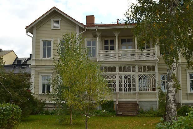 trähus i två våningar i beige liggande panel med vita knutar och detaljer. Balkong och veranda med mycket snickarglädje i vitt. Rött plåttak.