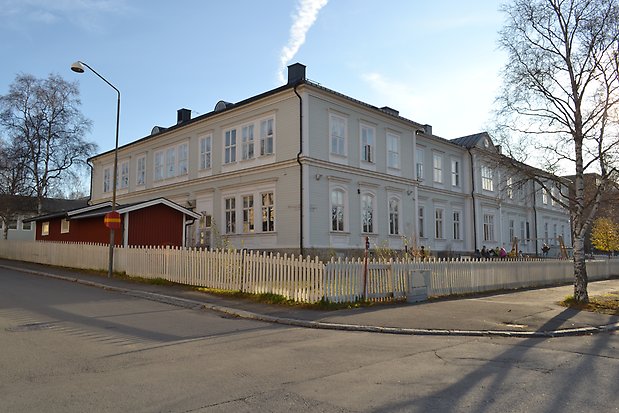 Skolbyggnad i två våningar i ljusgrå, liggande panel. Vita detaljer, Svart plåttak och hängrännor.