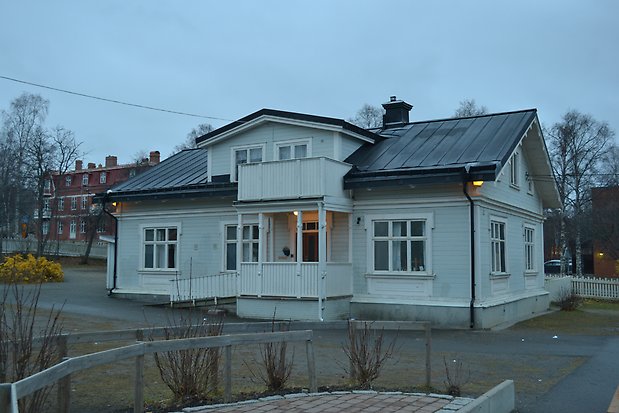 Villa i två våningar i ljusgrå, liggande panel. Vita knutar, detaljer och fönsterfoder, vita fönster med spröjs. Svart plåttak. Balkong och förstukvist med vitt staket.