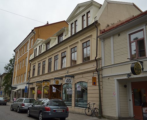 Stenbyggnad i tre våningar i ljusgul puts. Bruna fönster med spröjs. 