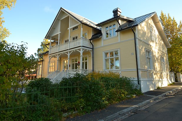 Villa i tre våningar, gul liggande panel med vita detaljer. Svart plåttak. Veranda och balkong med mycket vit snickarglädje. 