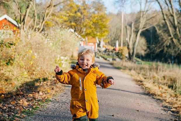 Glad pojke springer och skrattar på liten väg som kantas av färgglada höstlov. I bakgrunden skymtar röda hus.