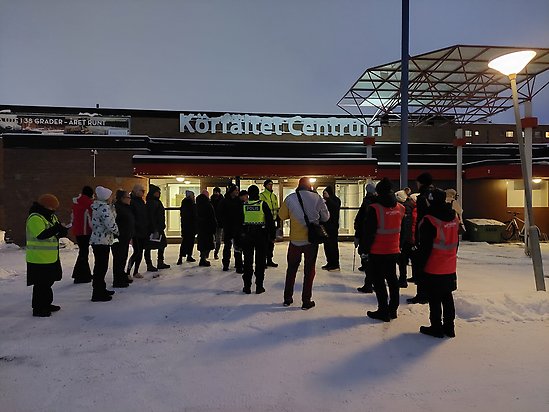 En grupp personer i vinterkläder, några med röda reflexvästar och en polis med gul reflexväst, står framför en tegelbyggnad som det står "Körfältet centrum" på.