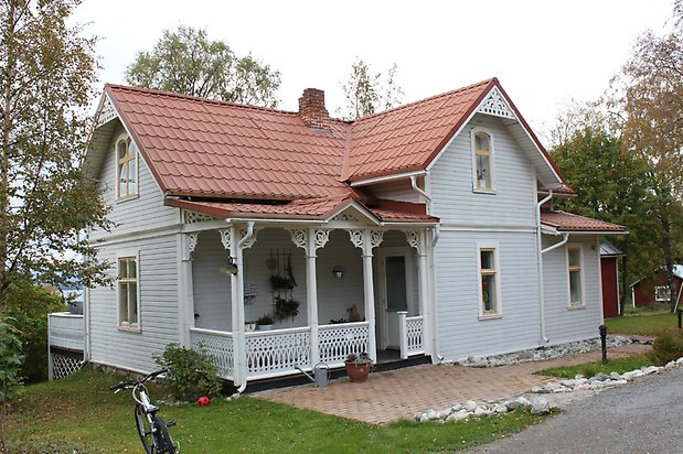 Spinetten 1. Villa i ljusgrå, liggande panel. Vita fönster med gula spröjs, veranda med snickarglädje och rött tegeltak.