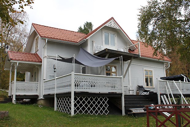 Spinetten 1. Villa i ljusgrå, liggande panel. Vita fönster med gula spröjs, veranda med snickarglädje och rött tegeltak.