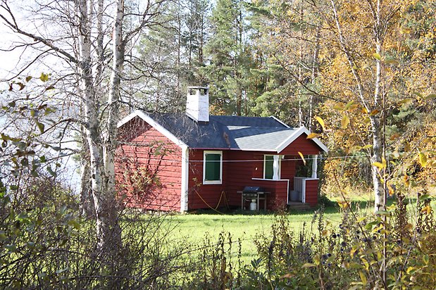 Kattfoten 1. Hus i röd, liggande panel, vita fönster med gröna luckor, grön dörr, och svart tak.