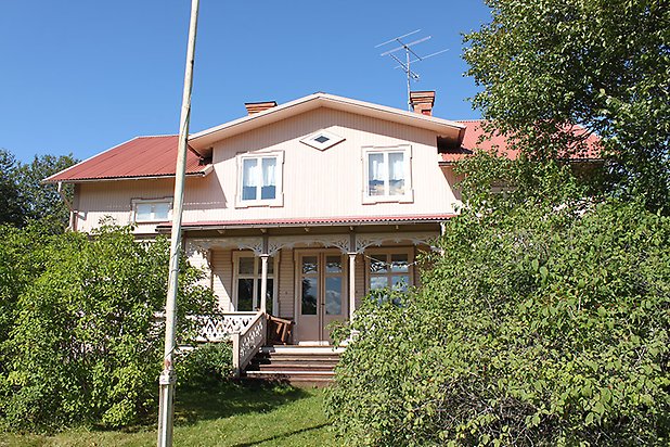 Söre 4:44. Villa i ljusbeige, liggande panel med mörkare detaljer och vita fönster med spröjs. Veranda med snickarglädje.