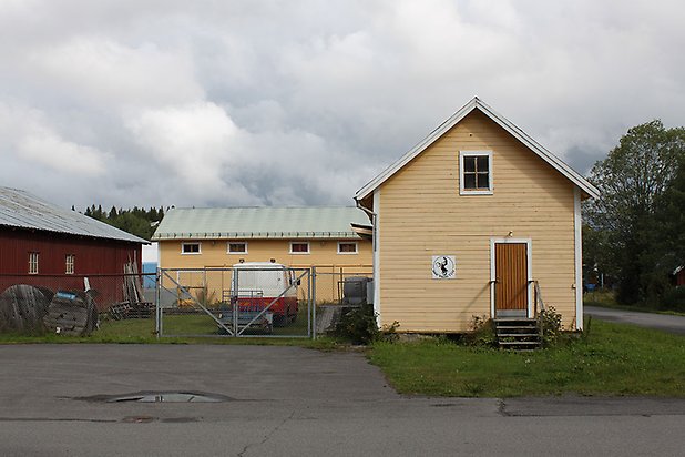 Söre 3:57. Byggnader i gul, liggande panel, brun dörr och små fönster.