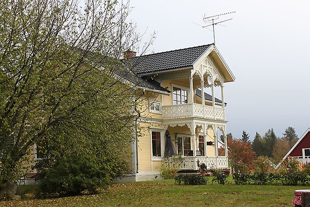 Söre 4:143. Villa i gul, liggande panel med vita fönster med röda spröjs och förstukvist och balkong med snickarglädje.