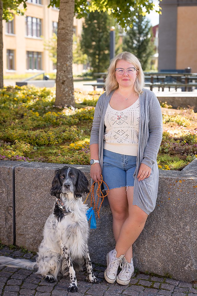 En medelålders kvinna med långt ljust hår och glasögon och en grå kofta stor med en svart och vit hund vid en stenmur