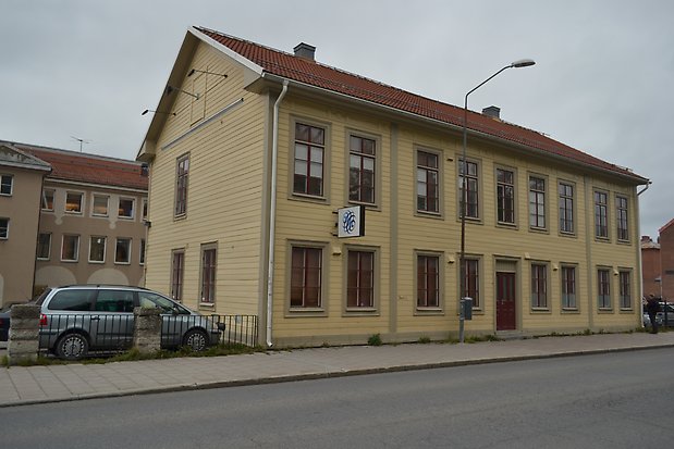 Trähus i två våningar i ljusgul liggande panel med ljusgröna detaljer, rött tak, bruna fönster med spröjs.