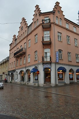 Stenbyggnad i fyra våningar i rosa puts med markplan i ljusgrå puts,. Vita fönster med spröjs, balkonger med svarta metalräcken. Skyltfönster i markplan.