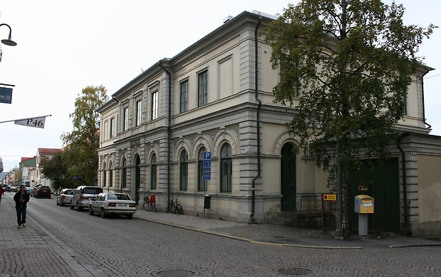 Stenbyggnad i två våningar. Ljusgrå puts. Fönster i valv på markplan. Svarta fönster med spröjs.