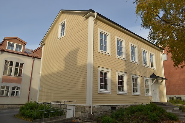 Trähus i tre våningar i ljusgul, liggande panel med vita detaljer, Vita fönster med spröjs, vit ytterdörr med litet svart tak över, stengrund och stenmur.