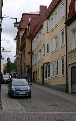 Stenbyggnad i tre våningar gul puts, vita fönster med spröjs, grå stengrund, burspråk på våning 2 och 3. Rött tak.