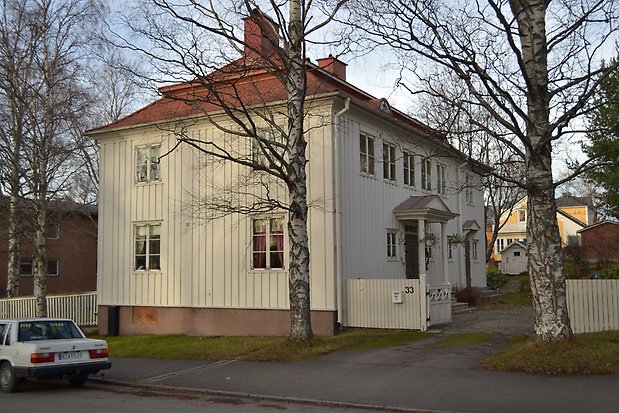 Trähus i två våningar i vit panel, vita fönster med spröjs, rött plåttak med röda skorstenar. Förstukvist med vita pelare.