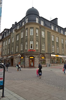 Stenbyggnad i fyra våningar i beige och mörkgrön puts, bottenplan i grått tegel. Skyltfönster i bottenplan. Vita fönster. Svart plåttak med takkupor och kupol.