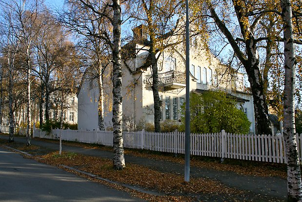 Stenvilla i tre våningar i vitputs. Svart plåttak. Balkong med svart räcke. Vita fönster med spröjs. Glasveranda.