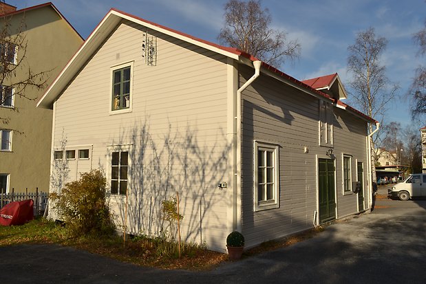 Trähus i två våningar i öjusgrå, liggande panel. Vita detaljer. Vita och gröna fönster med spröjs. Rött plåttak.