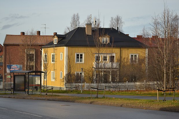 Trävilla i tre våningar i gul panel med vita detaljer. Svart plåttak med takkupor. Förstukvist med balkong över. 