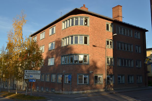 Brun tegelbyggnad i fyra våningar, rundat hörn med fönsterrad på våning 2-4. Blå fönster utan spröjs.