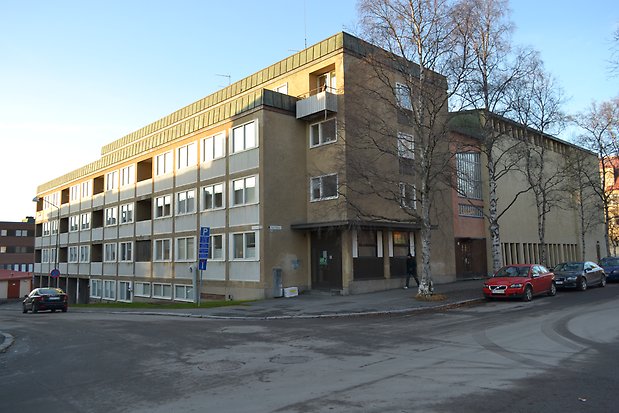 Byggnad i fyra våningar plus källarplan med garage. Beige puts, vita fönster med panelunder, balkonger inne i huskroppen. Svart port.