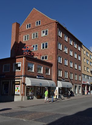 Brun tegelbyggnad i 2-5 våningar. Vita fönster utan spröjs, grå grund. Skyltfönster i markplan. Byggnaden har en del med 2 våningar med en röd neon-skylt.