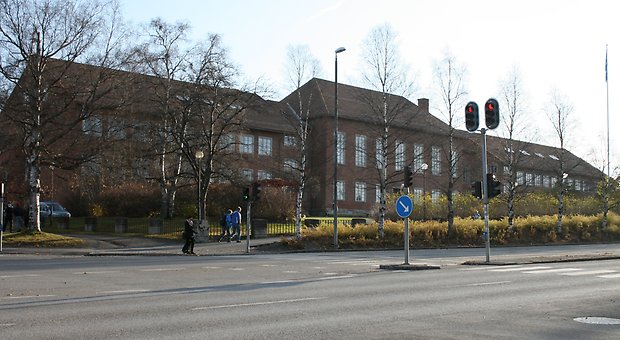 Brun tegelbyggnad i två våningar. Tre byggnadskroppar. Stora, vita fönster med spröjs. Brunt tegeltak.