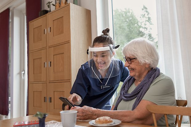 Yngre kvinna från hemtjänsten hjälper äldre kvinna med något på hennes mobiltelefon.