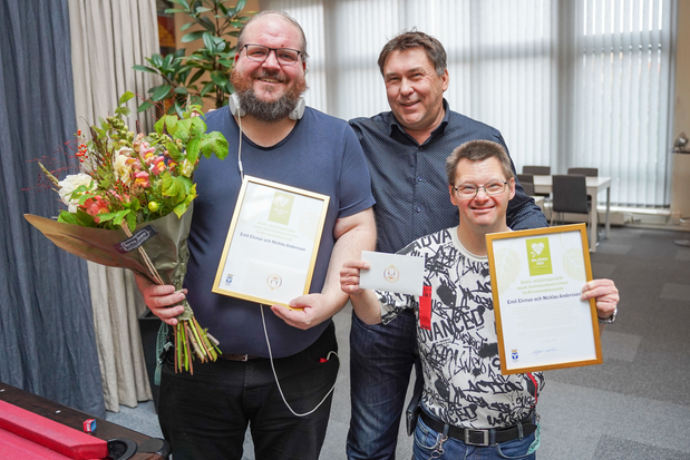 Tre män står bredvid varandra, mannen i mitten har armarna om männen till höger och vänster. Mannen till höger har ett diplom och en blombukett i händerna, mannen till höger har ett diplom och ett presentkort i händerna.